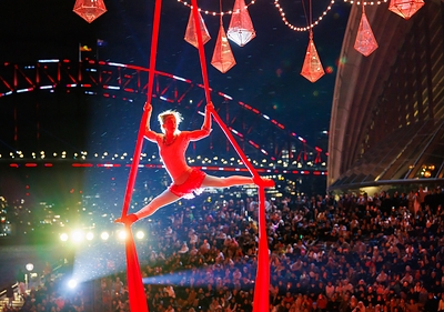 An aerial performer in a red costume executes a split on red silks during a Vivid Sydney 2026 event, with the Sydney Harbour Bridge and Opera House illuminated in the background and a large audience watching